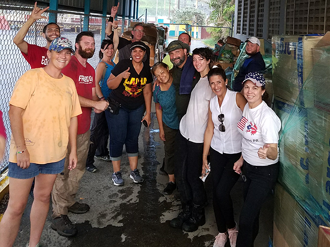Volunteers from seven different groups and organizations smile by pallets of supplies unloaded from the boat in Puerto Rico during Hurricane Maria response efforts.